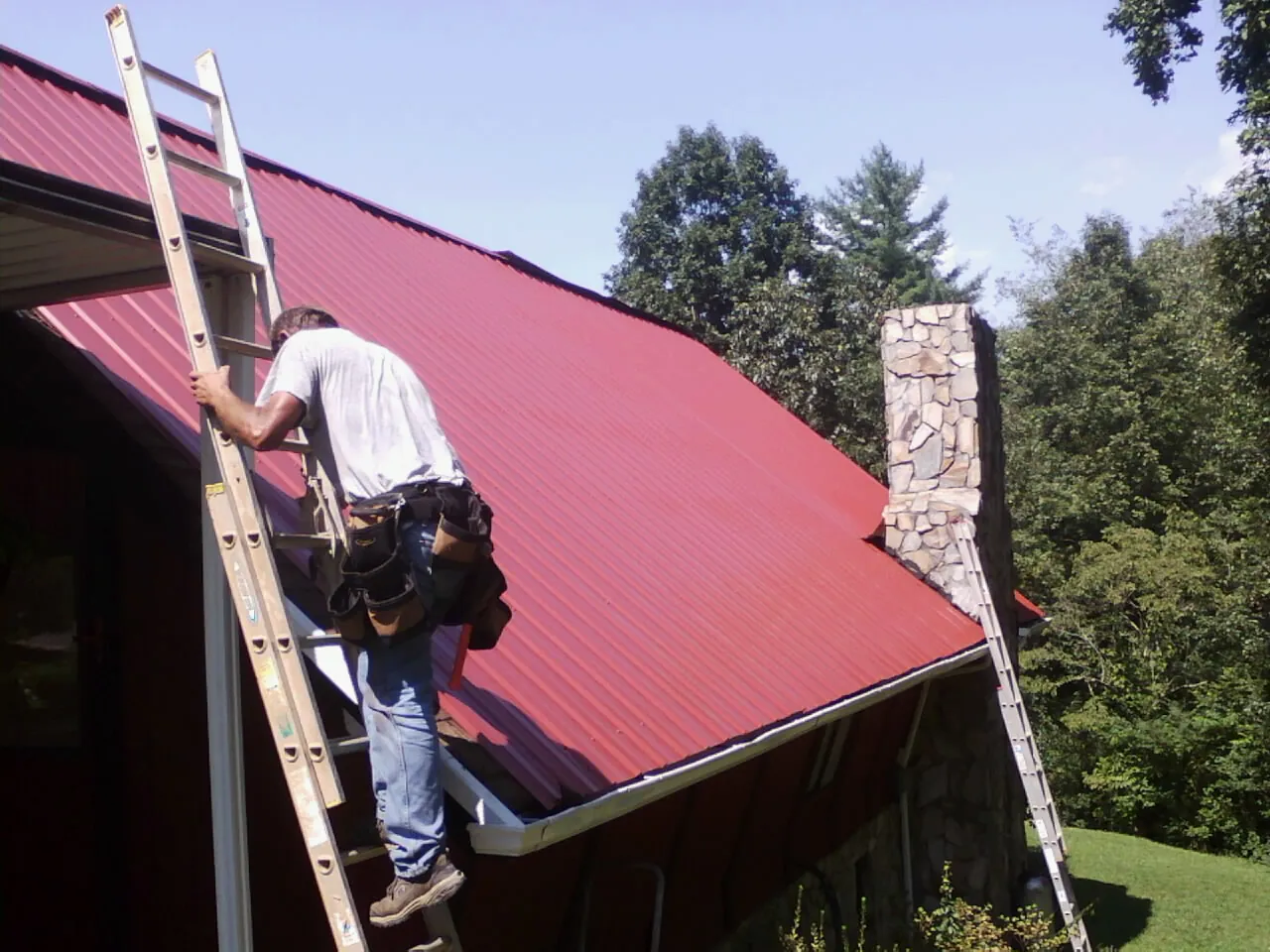 Roofer on a ladder beside a red metal roof and stone chimney — Soffit & Fascia Repair by Roofing Brothers in Ottsville